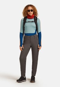 Frau mit lockigem Haar, die eine Sonnenbrille, ein langärmliges Shirt mit Blockfarben, graue Hose, Wanderschuhe, einen roten Schal und einen schwarzen Rucksack trägt.