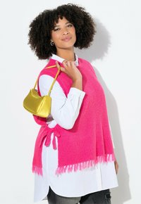 Heldroze gebreide poncho met franjes, gedragen over een witte button-up blouse, gecombineerd met een kleine, glanzende gele handtas.