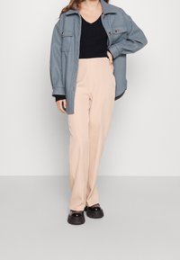 Giacca blu oversize con due tasche frontali, top nero, pantaloni beige su misura e scarpe chunky nere su uno sfondo semplice.