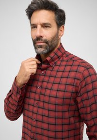 Camicia a quadri rossa con dettagli neri, colletto a camicia, e bottoni, caratterizzata da una texture morbida e un design aderente.