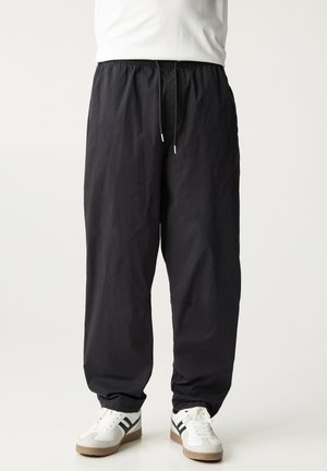 Personne portant un pantalon noir ample avec une taille élastique et des baskets blanches à rayures noires, debout devant un fond uni.