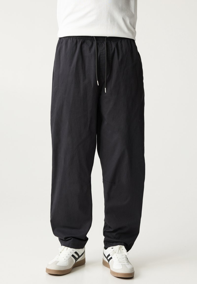 Personne portant un pantalon noir ample avec une taille élastique et des baskets blanches à rayures noires, debout devant un fond uni.