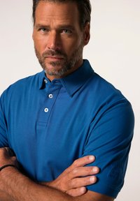 Blå polo skjorta med klassisk krage, tre knappar och korta ärmar. Har textuerad tyg och en logotypdetalj på ärmen.