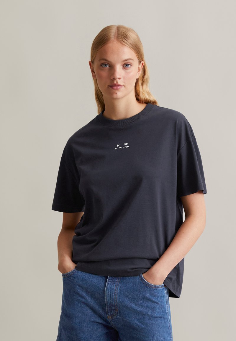 T-shirt en coton bleu marine à manches courtes, col rond, avec le texte blanc "Fais partie de mon histoire" imprimé sur le devant. Porté avec un jean bleu.