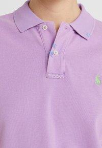 Levandulové polo tričko s texturovanou tkaninou, dvouknoflíkovou léga a vyšitým zeleným logem. Doplňuje jej světle modré prošívání kolem límce.