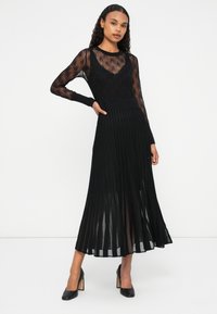 Vestido negro de manga larga con parte superior de texturizado y semitransparente, y falda larga plisada, complementado con zapatos de tacón alto negros.