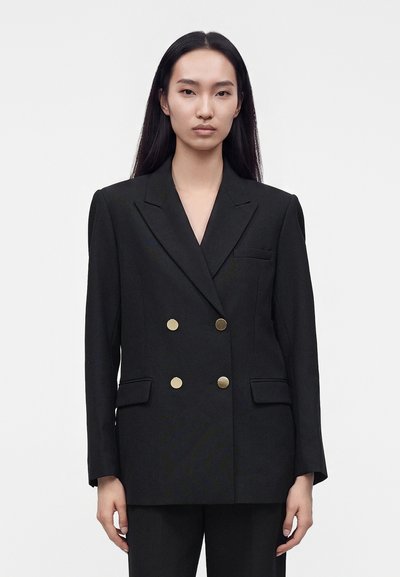 Personne aux longs cheveux noirs portant un blazer noir croisé avec des boutons dorés et un pantalon noir assorti sur un fond uni.