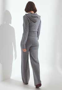 Donna con capelli corti castani che indossa un maglione grigio aderente con cappuccio e pantaloni coordinati a gamba larga, in piedi contro una parete bianca.