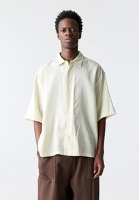 Chemise à manches courtes, boutonnée en jaune clair avec de subtiles rayures verticales. Fabriquée en tissu doux, elle présente un col pointu et une coupe décontractée.