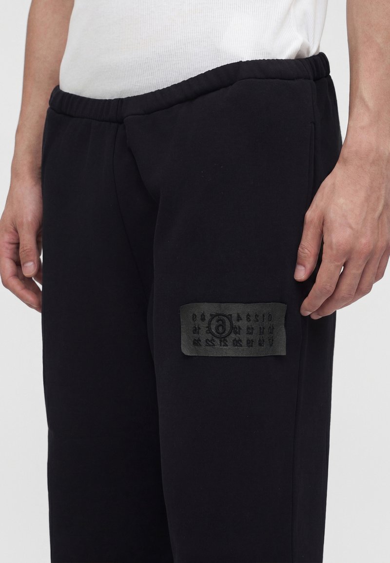 Gros plan d'une personne portant un pantalon de jogging noir avec une taille élastique et un patch noir avec des symboles sur la cuisse gauche.