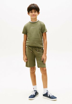 Jongen staat, draagt een olijfgroen shirt met korte mouwen en korte broek, witte sokken en marineblauwe sneakers tegen een effen witte achtergrond.