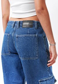 Pantalones de mezclilla azul con un ajuste relajado, que presentan una etiqueta negra en la cinturilla, dos bolsillos traseros y detalles de costura visibles.