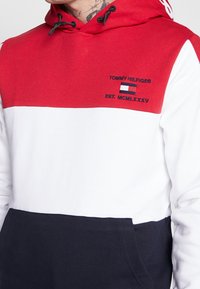 Sweat à capuche à blocs de couleur rouge, blanc et bleu marine avec le texte "Tommy Hilfiger EST. MCMLXXXV" et le logo du drapeau sur la poitrine.