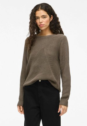 Brun strikket sweater med et tekstureret mønster, rund halsudskæring og lange ærmer. Kombineret med sorte bukser, der har en diskret frontknap.