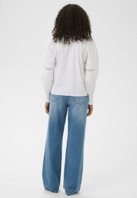 Femme aux cheveux bouclés portant un chemisier blanc à manches longues avec des détails en dentelle et un jean bleu à jambes larges, se tenant de dos sur un fond blanc.