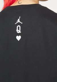Dos d’un t-shirt noir avec un logo Jordan jumpman blanc, une lettre Q stylisée et un symbole de cœur sous le cou d’une personne aux cheveux rouges avec un tatouage en forme de croissant.
