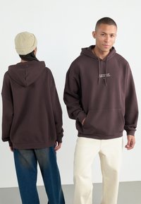 Brun hettegenser med lomme foran, med tekst på brystet. Kombinert med lyse, vide bukser og en beige beanie.