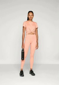 Világos rózsaszín sportos öltözet, csomózott crop toppal és testhez simuló leggings-szel. Az egyik kezében egy fekete vízpalackot tart.