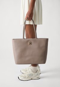 Sac fourre-tout en cuir gris avec deux poignées robustes et un accent en logo doré. Le sac présente une forme structurée et une texture lisse.