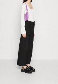 Žena v bílém dlouhém rukávu, černých širokých kalhotách, černých platformových mokasínech, se stříbrným náhrdelníkem a menší fialovou kabelkou přes rameno.