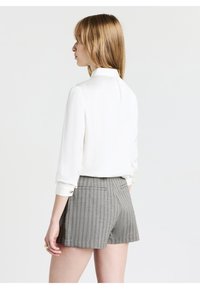 Blusa bianca con colletto alto, maniche lunghe e dettagli sui polsini; abbinata a pantaloni corti grigi a righe sottili, tagliati sopra la metà della coscia.