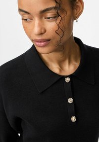 Žena s kovrčavom kosom koja nosi crnu košulju s ovratnikom i tri metalna teksturirana gumba, gleda prema dolje i u stranu.