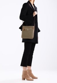 Olivengrøn crossbody taske med glat overflade, guldfarvet lynlås og brune detaljer. Båret med sorte bukser og en sort frakke.