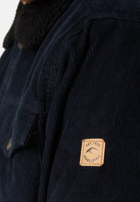 Marine corduroy jasje met een zwarte sherpa kraag. Heeft een borstzak en een tan gestikte label met "EST. 1993" en een logo.