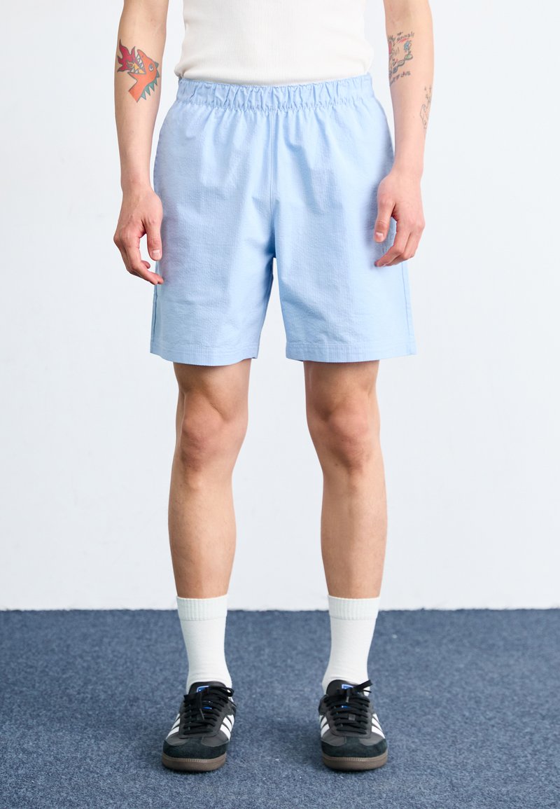 Shorts din bumbac de culoare albastru deschis cu talie elastică, buzunare laterale și o croială lejeră. Poartă șosete albe și pantofi sport negri.