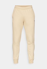 Beige sweatpants made from soft fabric, featuring a tapered leg, elastic waistband, side pockets, and a small logo on the left thigh.