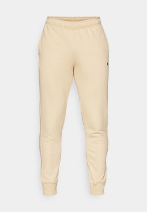 Pantalon de survêtement beige en tissu doux, avec une jambe fuselée, une taille élastique, des poches latérales et un petit logo sur la cuisse gauche.