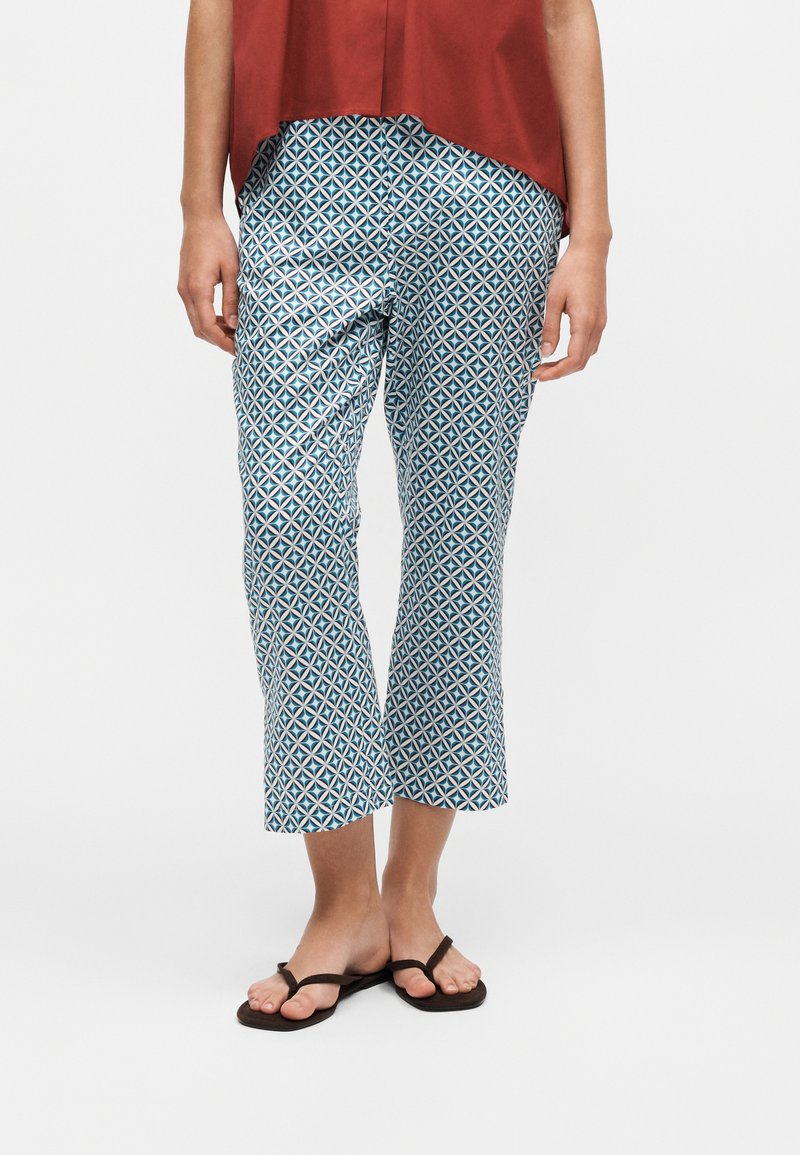Personne portant un pantalon court à motifs géométriques bleu et blanc, un haut couleur rouille et des tongs noires, debout sur un fond blanc.