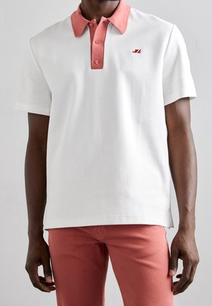 Man draagt een wit poloshirt met korte mouwen, een koraalkleurige kraag en knoopsluiting, gecombineerd met koraalkleurige broek, staand tegen een effen lichte achtergrond.