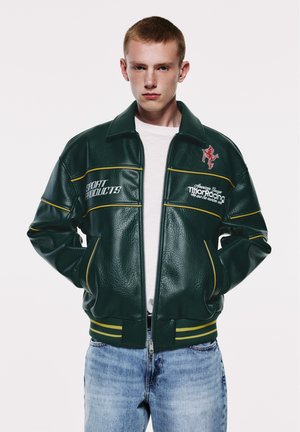 Jeune homme portant une veste en cuir vert foncé avec un passepoil jaune et un texte blanc, associé à un jean bleu clair, debout devant un fond uni.