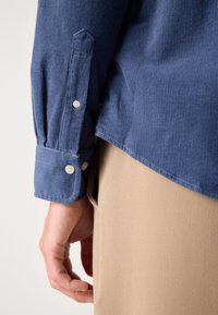 Gros plan d’une personne portant une chemise en velours côtelé bleue avec des poignets boutonnés et un pantalon beige, main détendue le long du corps.