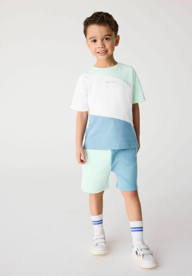 Detský outfit s tričkom s krátkym rukávom, ktoré má biele, svetlozelené a modré sekcie, v kombinácii s mentolovými šortkami. Doplnkové športové topánky.