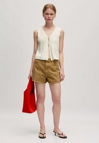 Witte, gestructureerde mouwloze top met een v-hals en een rits aan de voorkant, gecombineerd met khaki shorts en een rode draagtas. Zwarte sandalen maken de look compleet.