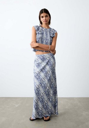 Žena nosiaca modro-biely vzorovaný bezrukávový crop top s volánikmi na ramenách a zodpovedajúcu dlhú sukňu, stojaca s prekríženými rukami.