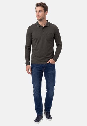 Mand iført mørkegrøn langærmet poloshirt, blå jeans og sorte sneakers, stående med den ene hånd i lommen og ser til venstre.