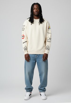 Sweatshirt crème avec graphiques imprimés en rouge, noir et bleu ; associé à un jean en denim bleu clair ; baskets blanches avec accents noirs.