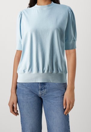 Kobieta ubrana w jasnoniebieską bluzkę z krótkim rękawem i bufkami na ramionach, w połączeniu ze średnio wypranymi niebieskimi dżinsami, stojąca na tle jednolitego tła.