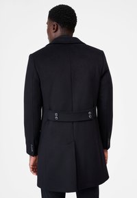 Svart ullkappa med skräddarsydd design, synliga knoppar på baksidan och midja med bälte. Har formella slag och strukturerade axlar.