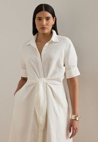 Balta lina krekla kleita ar pogām priekšpusē, īsām piedurknēm un sasienamu jostasvietu. Tai ir sānu kabatas un minimālas detaļas.