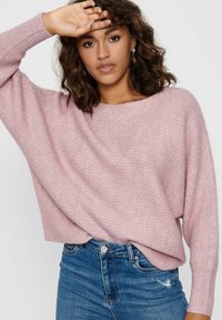 Pull en tricot surdimensionné rose clair avec une surface texturée et des manches larges, associé à un jean bleu délavé. Coupe décontractée avec un col rond.