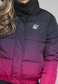 Puffa bunda v černé barvě, která přechází do růžové. Vybavena vysokým límcem, předním zipem a vyšitým logem v bílé barvě. Prošívaná textura s elastickým lemem.