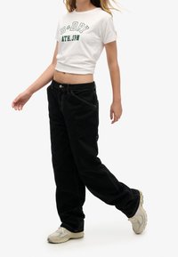 Camiseta blanca corta con texto verde, combinada con pantalones holgados negros y zapatillas de color claro. Diseño sencillo con detalles de costura visibles.