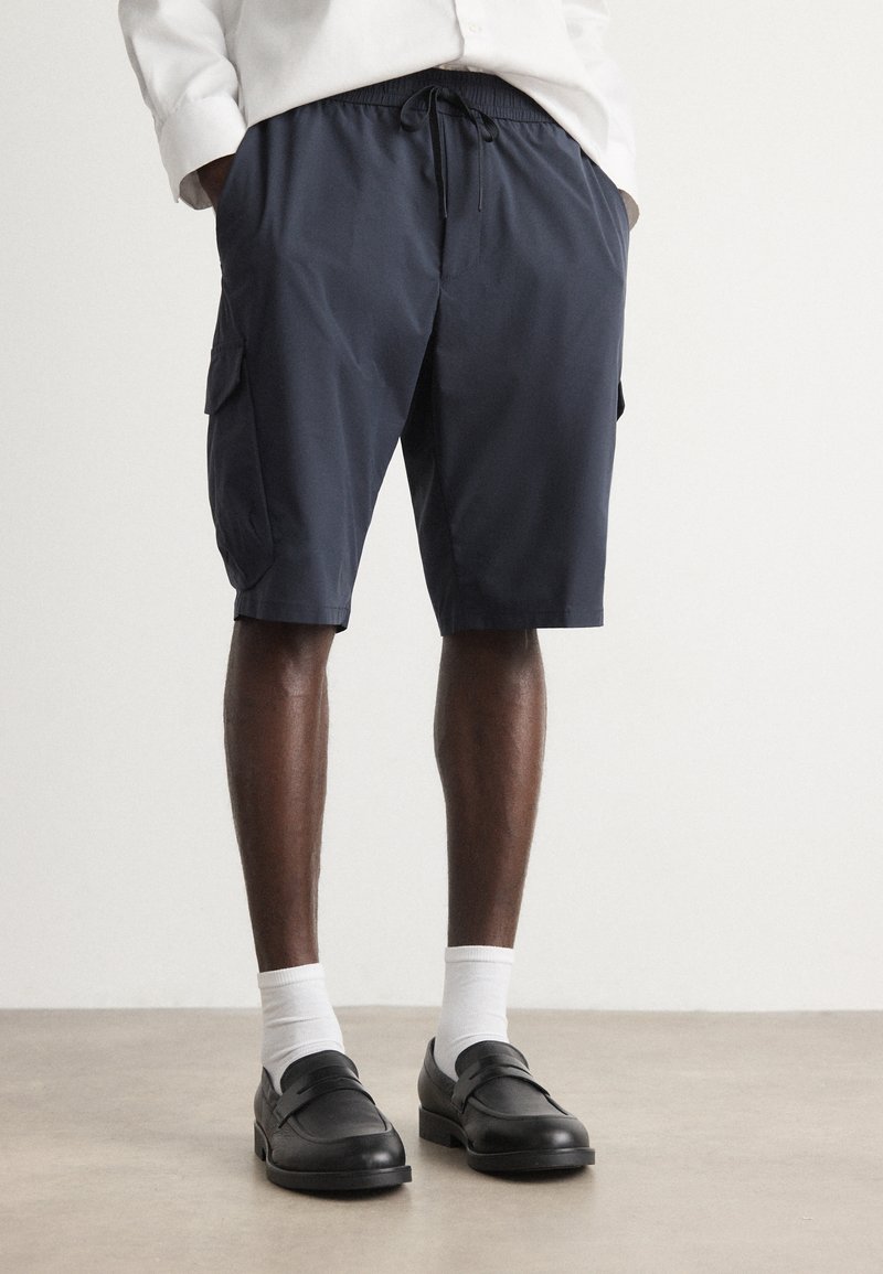 Shorts de cargo bleu marine en matériau léger, dotés de poches latérales et d'une taille élastique, associés à des mocassins noirs et des chaussettes blanches.
