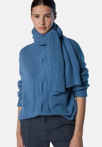 Maglione a treccia e sciarpa coordinata in una morbida tonalità di blu, che mette in risalto motivi testurizzati e una vestibilità rilassata. Il tessuto appare caldo e accogliente.