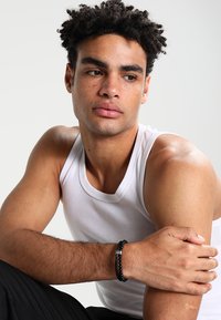 Black beaded bracelet with silver hardware on a male model's wrist. Model wears a white tank top, seated with arms crossed. Smooth skin texture.
