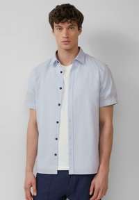 Himmelblaues Kurzarmhemd mit Knopfleiste und schwarzen Knöpfen. Mit klassischem Kragen und strukturiertem Stoff. Über einem weißen Hemd getragen.